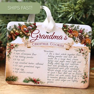 May include: A rectangular ceramic ornament featuring a white ribbon hanger and a printed recipe for Grandma's Christmas Cookies. The design includes festive illustrations of pine branches, red berries, pinecones, and dried orange slices.