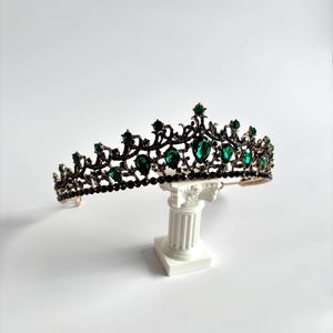 May include: A black and gold tiara with green gemstones. The tiara is on a white pedestal.