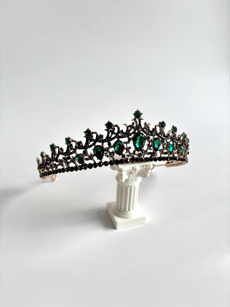 May include: A black and gold tiara with green gemstones. The tiara is on a white pedestal.