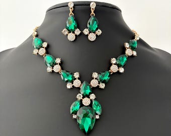 May include: A gold-toned necklace and earring set featuring large, emerald green teardrop-shaped gemstones. The necklace has a central pendant and smaller stones along the chain. The earrings mirror the necklace design with matching green and clear stones.