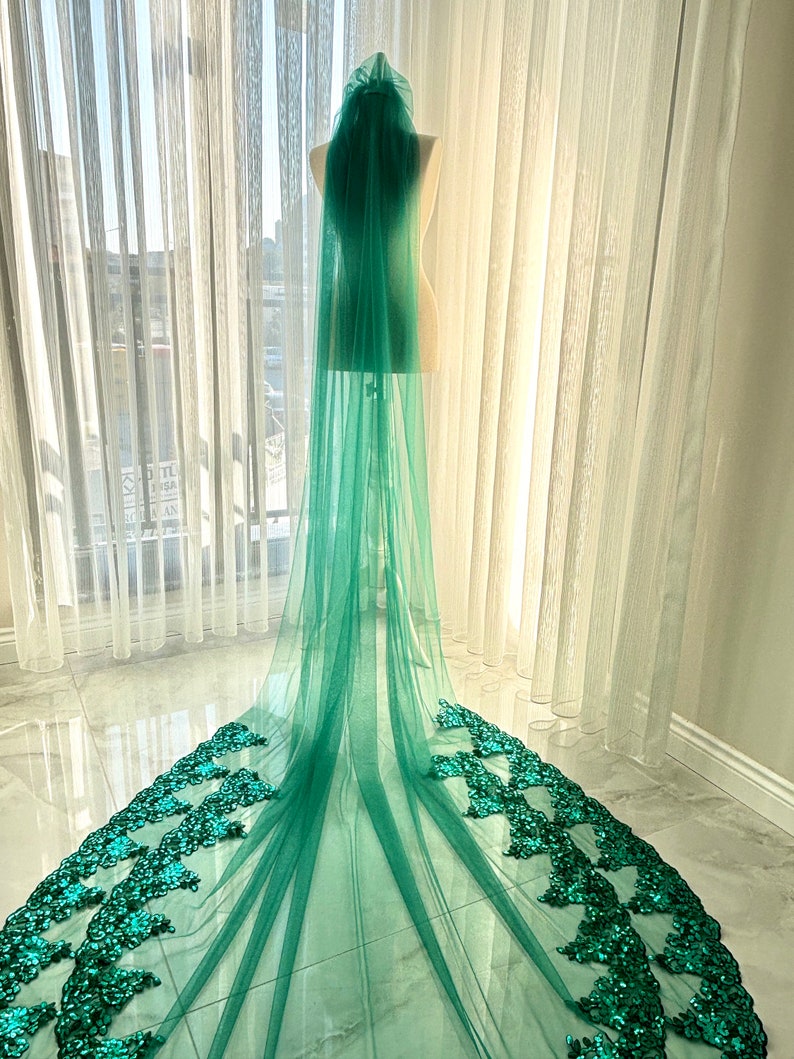 May include: A long, green tulle wedding veil with a sequined border. The veil is draped over a mannequin and is displayed against a white background.