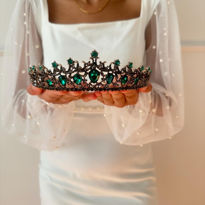 May include: A gold and green tiara with emerald green gemstones. The tiara is held in the hands of a person wearing a white dress.