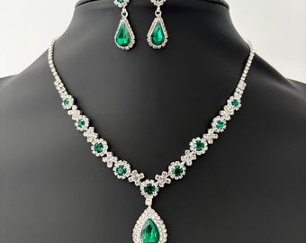 May include: A silver necklace and earring set with emerald green teardrop gemstones. The necklace has a central pendant and matching drop earrings, all set with clear crystals. A classic jewellery set.