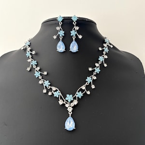 Puede incluir: Un conjunto de collar y pendientes de plata con un colgante de lágrima y detalles florales. La joyería presenta cristales de color azul claro.