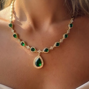 May include: A gold necklace with a large emerald green teardrop pendant surrounded by smaller clear stones. The necklace is made up of a chain with smaller emerald green teardrop stones and clear stones.