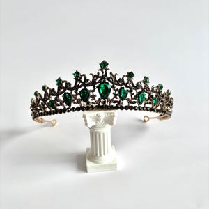 May include: A gold tiara with a black metal base and green gemstone accents. The tiara is on a white pedestal.
