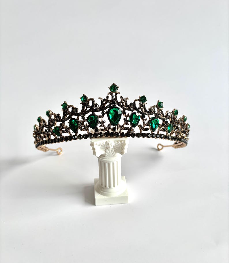 May include: A gold tiara with a black metal base and green gemstone accents. The tiara is on a white pedestal.