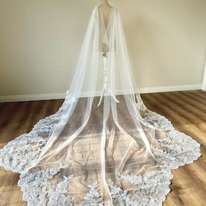 以下が含まれることがあります： 繊細なレースのトリムが付いた白いチュールのウェディングベール。ベールは長く、流れるような、チャペルトレインです。