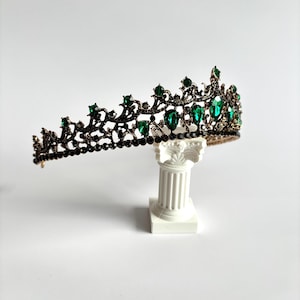 May include: A black crown with green gemstones. The crown is on a white pedestal.