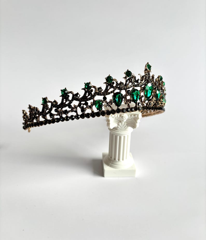 May include: A black crown with green gemstones. The crown is on a white pedestal.
