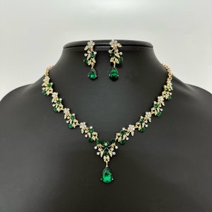 Può includere: Un set di collana e orecchini in oro con pietre di cristallo verdi e trasparenti. La collana presenta un ciondolo a forma di lacrima e un delicato motivo floreale.