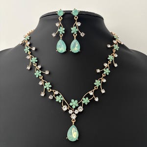 以下が含まれることがあります： 繊細な花のデザインと涙型の緑色の石がついた、金のネックレスとイヤリングのセットです。