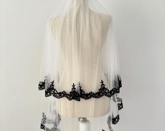 May include: A white bridal veil with two layers of black lace trim. The veil is displayed on a white mannequin.