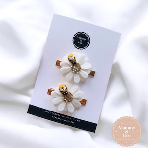 May include: Two white felt daisy hair clips with a yellow and black bee charm on each. The clips are on a white background with the text "Mommy & Gee Handmade with Love" and a logo.