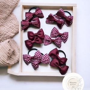 May include: A collection of burgundy and white gingham hair bows with matching solid burgundy bows. The bows are arranged in a wooden tray with a white circle sticker that says "Hair Accessories Mummy & Gee".