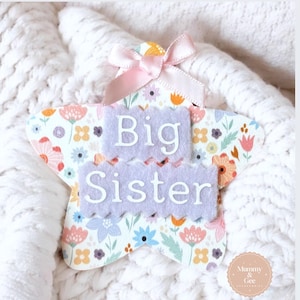 May include: A white star-shaped wooden sign with a floral pattern. The sign has two purple felt pieces with the words "Big Sister" written in white.