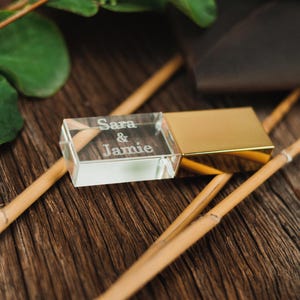 Può includere: Un'unità USB in cristallo trasparente con un rivestimento in metallo dorato. L'unità è incisa con i nomi "Sara & Jamie".