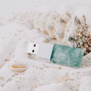 以下が含まれることがあります： 白い砂浜に貝殻とヒトデが置かれた、白と緑の貝殻模様のUSBメモリです。