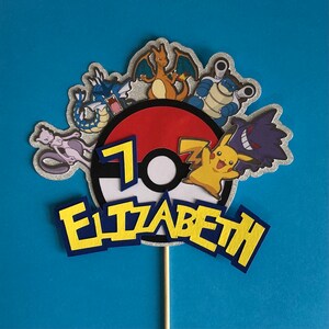 May include: A cake topper featuring a red and white Pokeball with a number 7, surrounded by various Pokemon characters including Pikachu, Charizard, and a Ghost-type Pokemon. The topper also includes the name "Elizabeth" in yellow letters with blue outlines.
