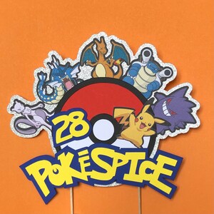 May include: A cake topper featuring a Pok&eacute; Ball with the number 28, surrounded by various Pok&eacute;mon characters, including Pikachu, Charizard, Blastoise, and Snorlax. The text "Pok&eacute;Spice" is written in a bold, yellow font.