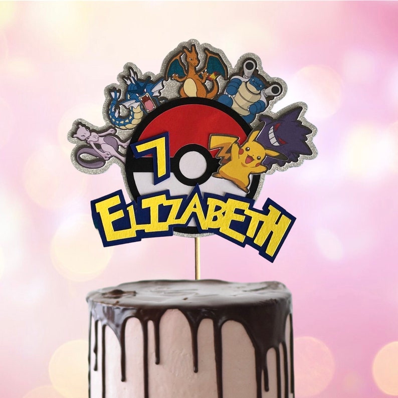 May include: A cake topper featuring a red and white Pokeball with the number 7, surrounded by various Pokemon characters, including Pikachu, Charizard, and Mewtwo. The topper also includes the name "Elizabeth" in yellow letters with blue outlines.