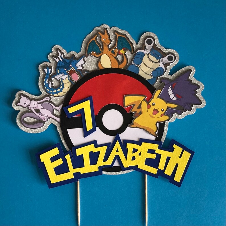 May include: A cake topper featuring a red and white Pokeball with a number 7, surrounded by various Pokemon characters including Pikachu, Charizard, Blastoise, and a purple and black Ghost-type Pokemon. The name Elizabeth is written in blue and yellow letters.