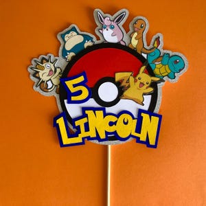 May include: A red and white Pokeball cake topper featuring a yellow Pikachu, a blue Squirtle, a pink Jigglypuff, a brown Eevee, and a blue and green Snorlax. The topper also includes the number 5 and the name Lincoln in yellow letters with blue outlines.