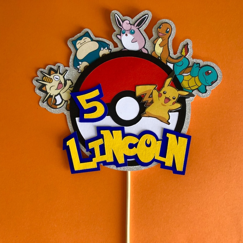 May include: A red and white Pokeball cake topper featuring a yellow Pikachu, a blue Squirtle, a pink Jigglypuff, a brown Eevee, and a blue and green Snorlax. The topper also includes the number 5 and the name Lincoln in yellow letters with blue outlines.