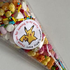 May include: A clear plastic cone filled with colorful candy and sprinkles. A white circle sticker with a cartoon Pikachu and the text "Thank you for coming to my party Love from Grayson" is attached to the cone.