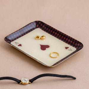 May include: A rectangular tray featuring an ace of hearts playing card design with a dark red and black checkered border. The tray holds two small gold hoop earrings and a thin gold ring. A wristwatch with a black strap lies below the tray.