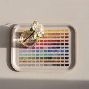 May include: A rectangular off-white serving tray featuring a grid of numerous small, rectangular color swatches in a spectrum of hues, each labeled with text. A small, clear glass vase containing a single white flower sits on the tray.