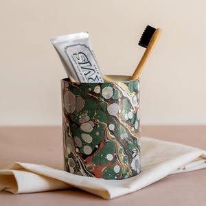 May include: A cylindrical bathroom tumbler featuring a marbled pattern in shades of dark green, rust orange, and grey. Inside the tumbler sits a tube of Marvis toothpaste and a bamboo toothbrush with black bristles, creating a functional set.