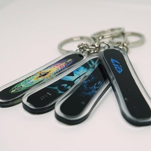 May include: Four snowboard-shaped keychains with different designs. One keychain is black with the word "LIB" in blue lettering. The other three keychains have colorful abstract designs.