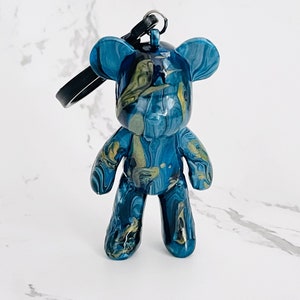Pode incluir: Uma estatueta de urso de resina marmorizada azul e dourada com uma al&ccedil;a preta. O urso tem um acabamento brilhante e um acess&oacute;rio de chaveiro. A estatueta &eacute; um item decorativo.