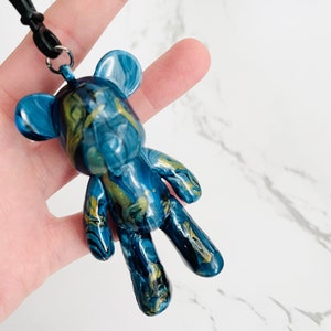 Pode incluir: Pingente de urso de pel&uacute;cia azul e dourado marmorizado com um cord&atilde;o preto. O urso tem um acabamento brilhante e um padr&atilde;o &uacute;nico. O pingente &eacute; um acess&oacute;rio decorativo.