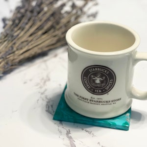 Peut inclure: Une tasse en c&eacute;ramique blanche avec un logo Starbucks brun et du texte qui dit "Starbucks Coffee Tea Spices Est. 1971 The First Starbucks Store Pike Place Market Seattle, WA". La tasse est pos&eacute;e sur un dessous-de-verre bleu turquoise.