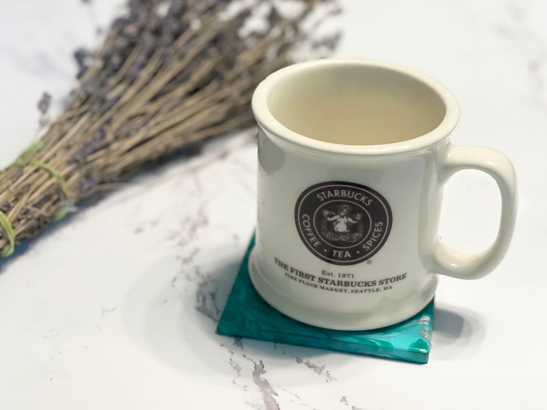 Peut inclure: Une tasse en c&eacute;ramique blanche avec un logo Starbucks brun et du texte qui dit "Starbucks Coffee Tea Spices Est. 1971 The First Starbucks Store Pike Place Market Seattle, WA". La tasse est pos&eacute;e sur un dessous-de-verre bleu turquoise.