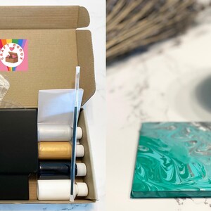 May include: A craft box with a rainbow and heart logo that says "Craft Goodie Box". The box contains a black canvas, paint bottles, a plastic stirring stick, and plastic gloves. The box is open and the contents are visible. A finished teal and silver abstract painting is shown on a white surface.