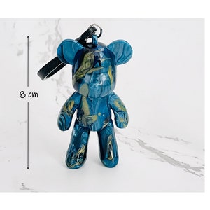 Pode incluir: Uma estatueta de urso em resina azul e dourada, com aproximadamente 8 cm de altura, com uma al&ccedil;a preta. O urso tem um design marmorizado com redemoinhos de ouro e azul. O objeto &eacute; decorativo.
