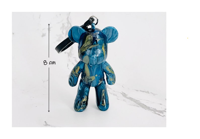 Pode incluir: Uma estatueta de urso em resina azul e dourada, com aproximadamente 8 cm de altura, com uma al&ccedil;a preta. O urso tem um design marmorizado com redemoinhos de ouro e azul. O objeto &eacute; decorativo.