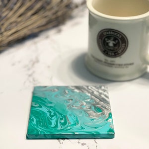 May include: A square coaster with a teal, white and silver marbled design. The coaster is made of a smooth, hard material and has a slightly raised edge.