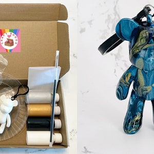 Pode incluir: Um kit de caixa de artesanato com um adesivo com o logotipo do arco-&iacute;ris, contendo uma luva de pl&aacute;stico transparente, um chaveiro em forma de urso branco e v&aacute;rios materiais de artesanato. Um chaveiro em forma de urso azul e dourado com uma al&ccedil;a preta tamb&eacute;m &eacute; mostrado.