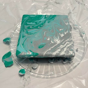 May include: A square coaster with a marbled design in shades of teal, grey, and white. The coaster is sitting on a clear plastic plate.