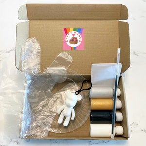 Pode incluir: Uma caixa de papel&atilde;o aberta contendo materiais de artesanato. Dentro est&atilde;o carret&eacute;is de linha nas cores branca, dourada, preta e branca, um pincel, um modelo de pl&aacute;stico transparente e uma estatueta de urso branco. Um adesivo com um arco-&iacute;ris e uma caixa em forma de cora&ccedil;&atilde;o est&aacute; na caixa.