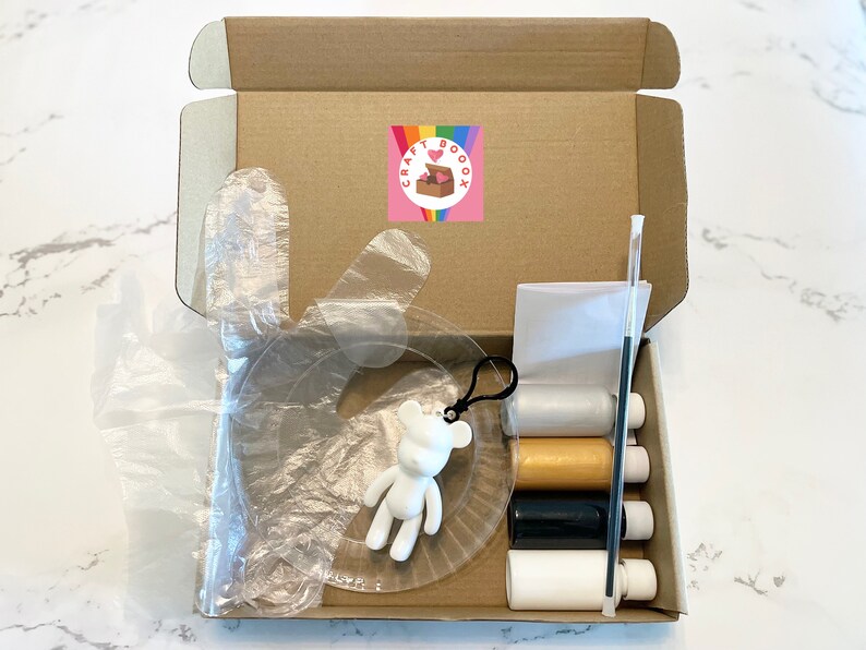 Pode incluir: Uma caixa de papel&atilde;o aberta contendo materiais de artesanato. Dentro est&atilde;o carret&eacute;is de linha nas cores branca, dourada, preta e branca, um pincel, um modelo de pl&aacute;stico transparente e uma estatueta de urso branco. Um adesivo com um arco-&iacute;ris e uma caixa em forma de cora&ccedil;&atilde;o est&aacute; na caixa.