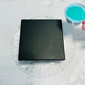 Peut inclure: Une tuile carr&eacute;e noire est pos&eacute;e sur une assiette en plastique transparente avec un motif en spirale. Une petite tasse en plastique avec un tourbillon de peinture bleu turquoise est tenue dans une main.