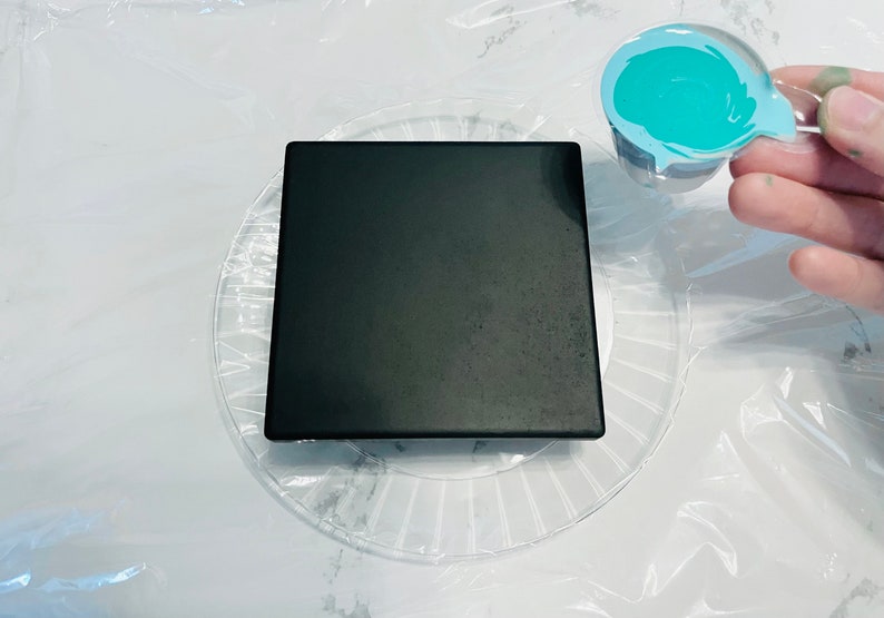 Peut inclure: Une tuile carr&eacute;e noire est pos&eacute;e sur une assiette en plastique transparente avec un motif en spirale. Une petite tasse en plastique avec un tourbillon de peinture bleu turquoise est tenue dans une main.