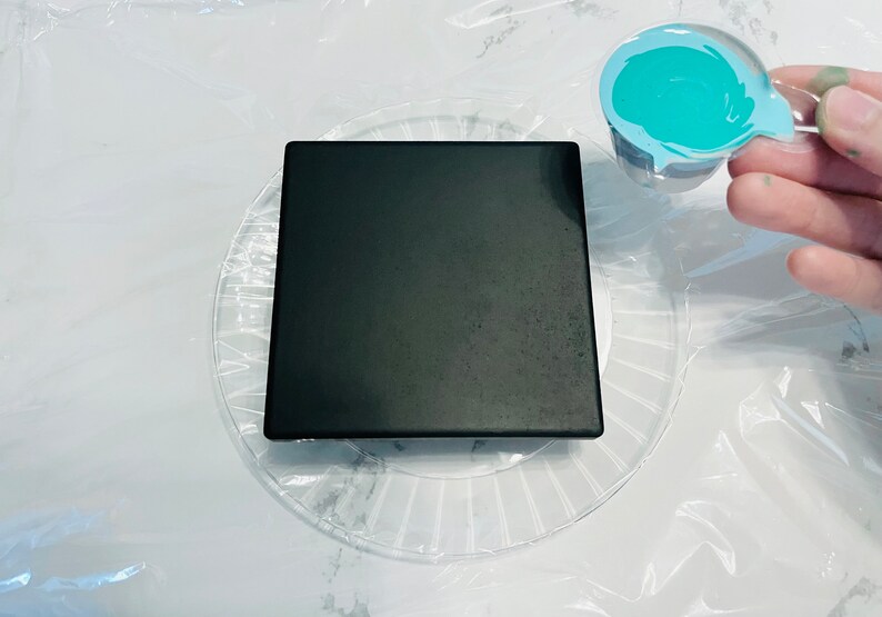 May include: A black square tile sits on a clear plastic plate with a swirling pattern. A small plastic cup with a teal blue paint swirl is being held above the tile.