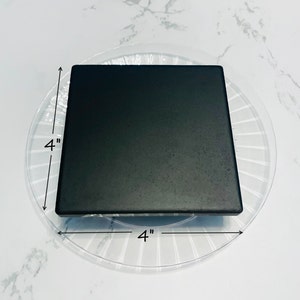 May include: A black square tile measuring 4 inches by 4 inches. The tile is sitting on a clear plastic plate with a decorative pattern.