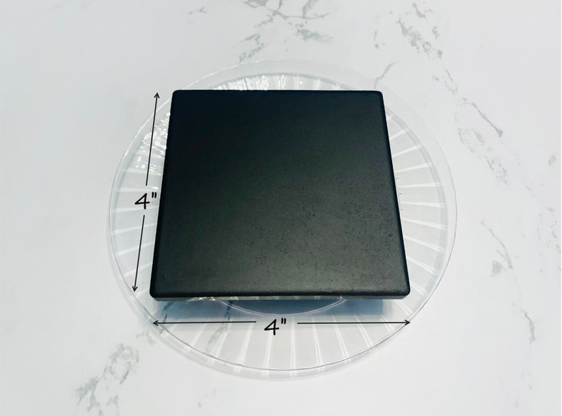 May include: A black square tile measuring 4 inches by 4 inches. The tile is sitting on a clear plastic plate with a decorative pattern.